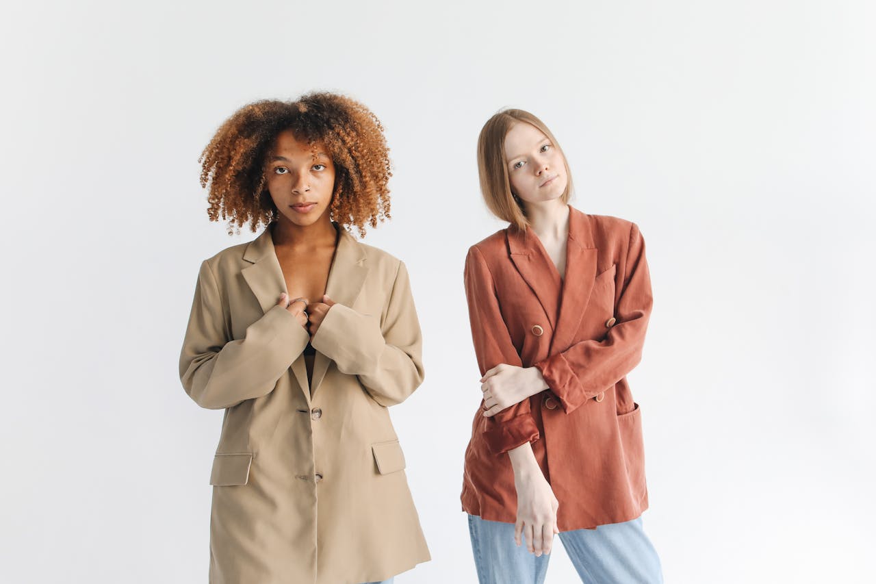 about-img-02 Two women modeling fashionable blazers in a studio setting with a white backdrop.